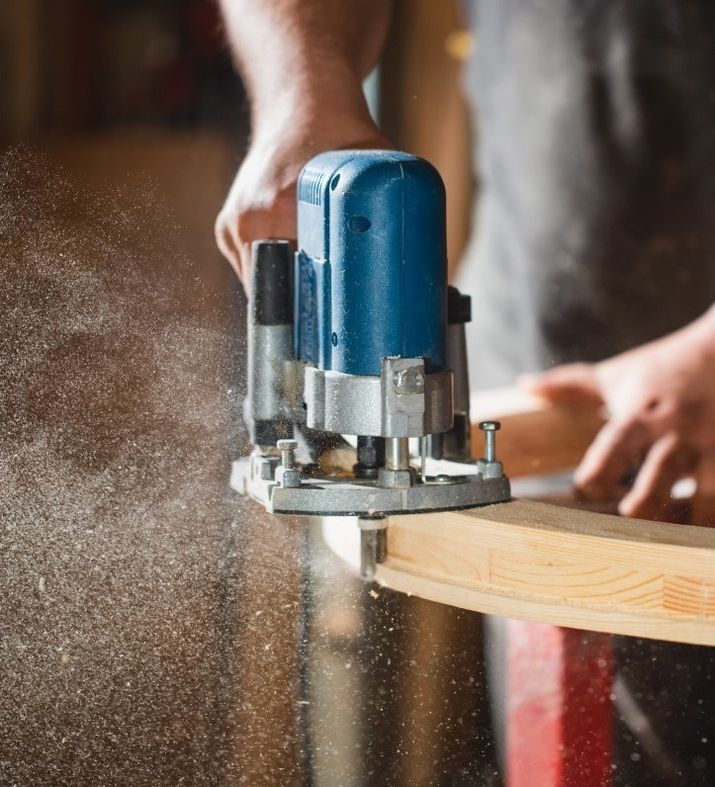 Handwerker fräst eine Holzleiste mit einer blauen Oberfräse, Staubpartikel fliegen durch die Luft