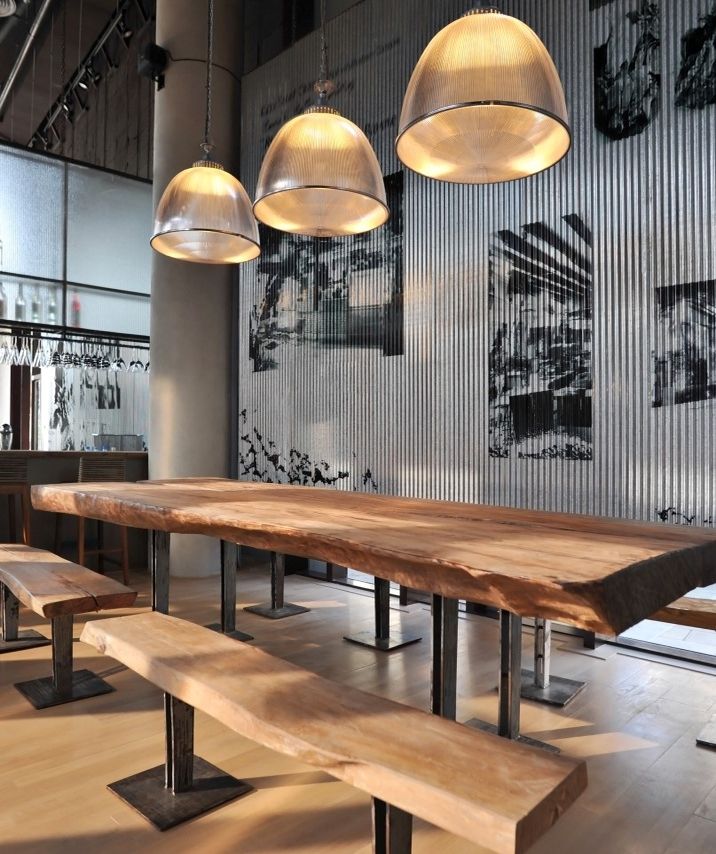 Restaurant im industriellen Stil mit einem großen, massiven Holztisch und Bänken, beleuchtet von Hängelampen.
