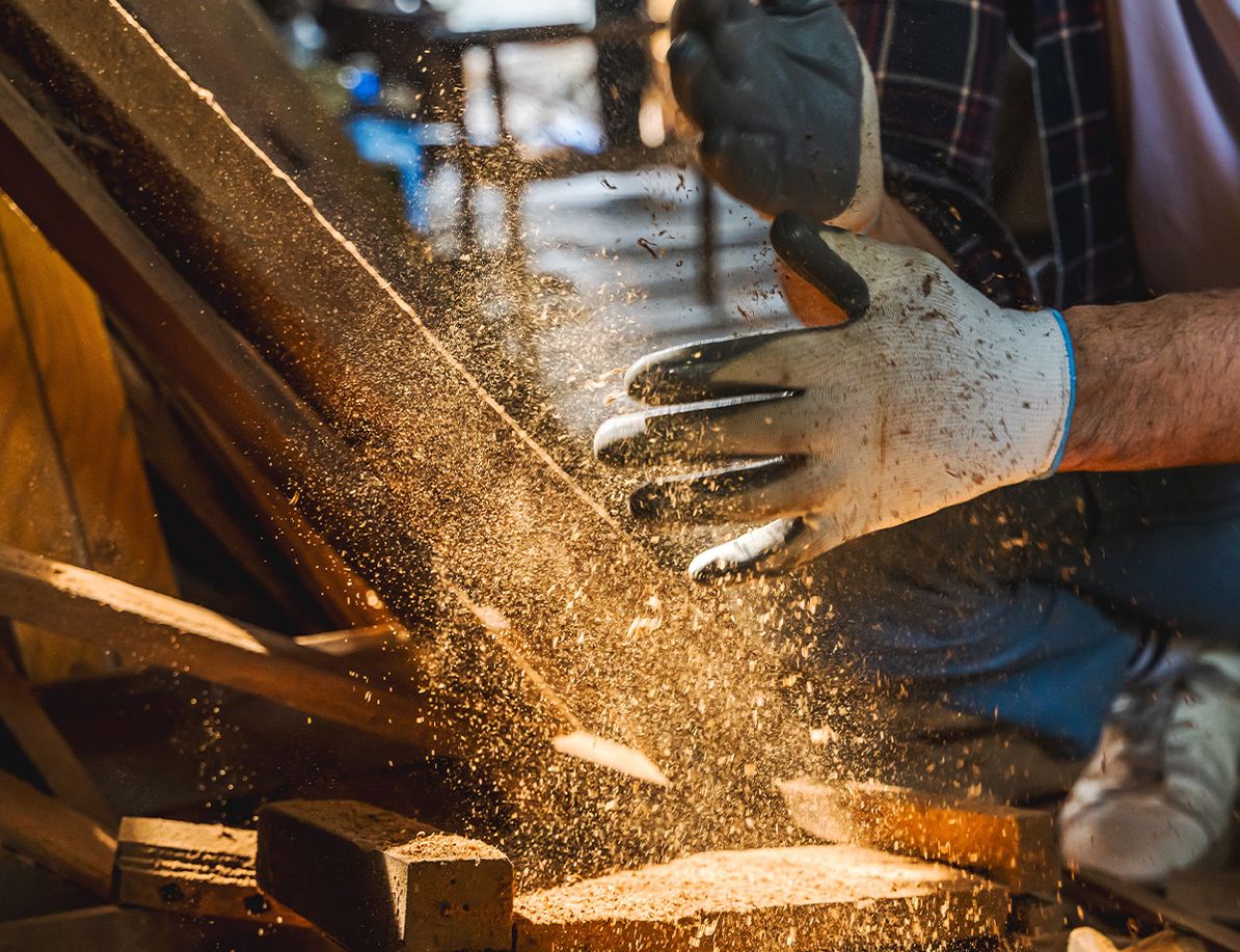 Handwerker mit Schutzhandschuhen bearbeitet Holz, während Staub und Späne in die Luft wirbeln.